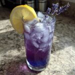 A tall glass filled with bubbly, purple Iced Blueberry Lavender Lemonade, ice, and garnished with a lemon slice and lavender sprigs.