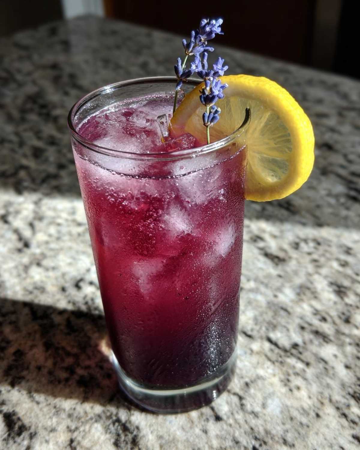 A tall glass filled with vibrant Iced Blueberry Lavender Lemonade, ice, and garnished with lemon and lavender sprigs.