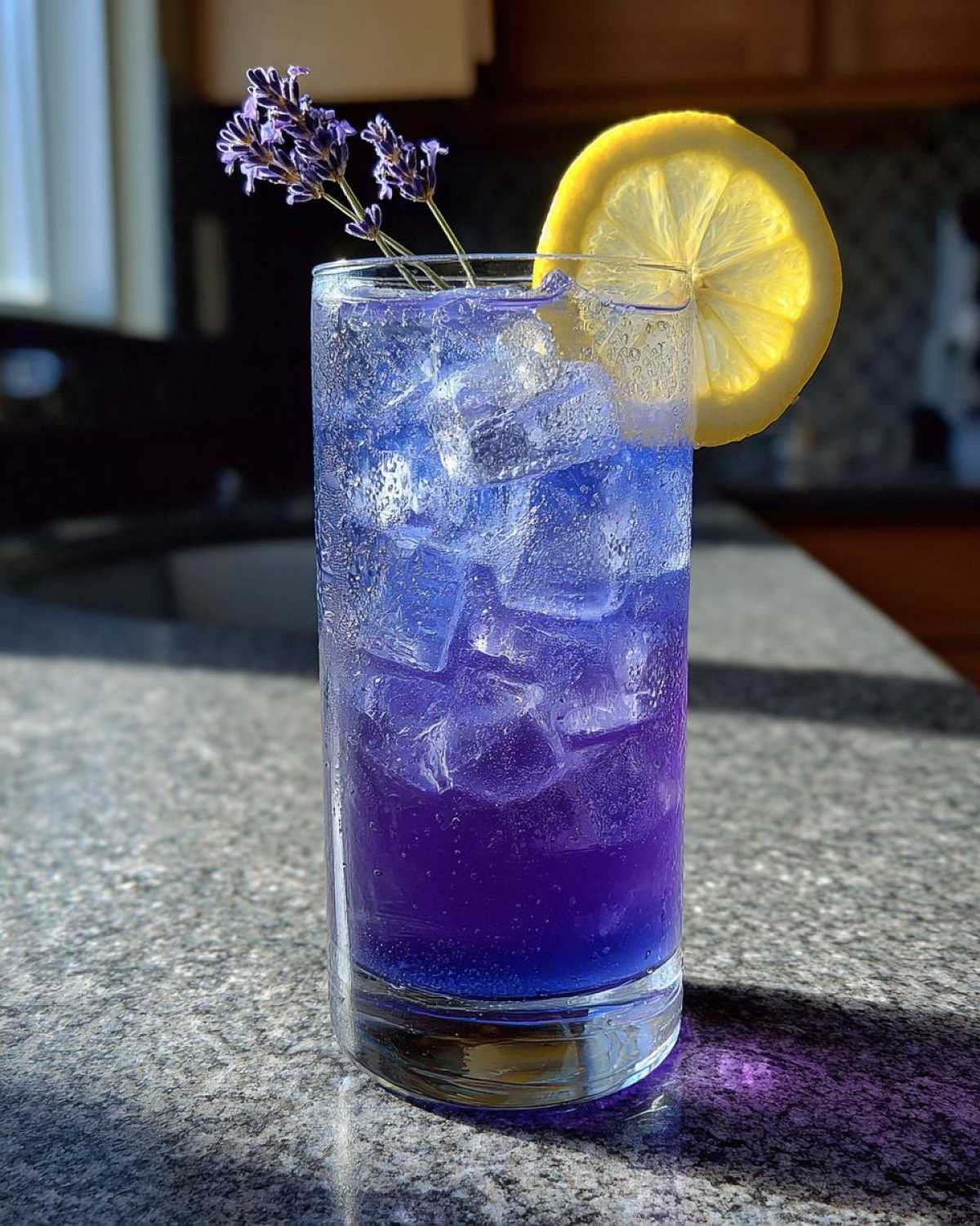 A tall glass filled with vibrant purple Iced Blueberry Lavender Lemonade, ice, and garnished with lavender sprigs and a lemon slice.