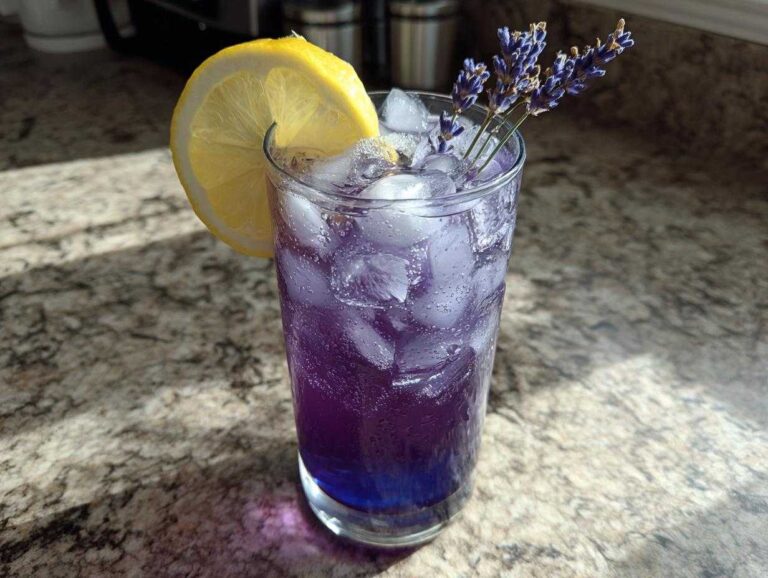 A tall glass filled with bubbly, purple Iced Blueberry Lavender Lemonade, ice, and garnished with a lemon slice and lavender sprigs.