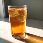 A tall glass filled with amber-colored Iced Peach Green Tea and ice cubes, sitting on a white counter in bright sunlight.