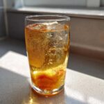A tall glass filled with bubbly Iced Peach Green Tea, ice cubes, and a slice of peach, sitting on a counter.