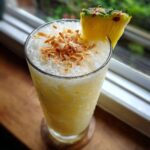 A tall glass of Iced Pineapple Coconut Mocktail garnished with toasted coconut flakes and a pineapple wedge.