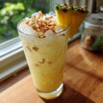 A tall glass filled with an Iced Pineapple Coconut Mocktail, topped with crushed ice and toasted coconut flakes.