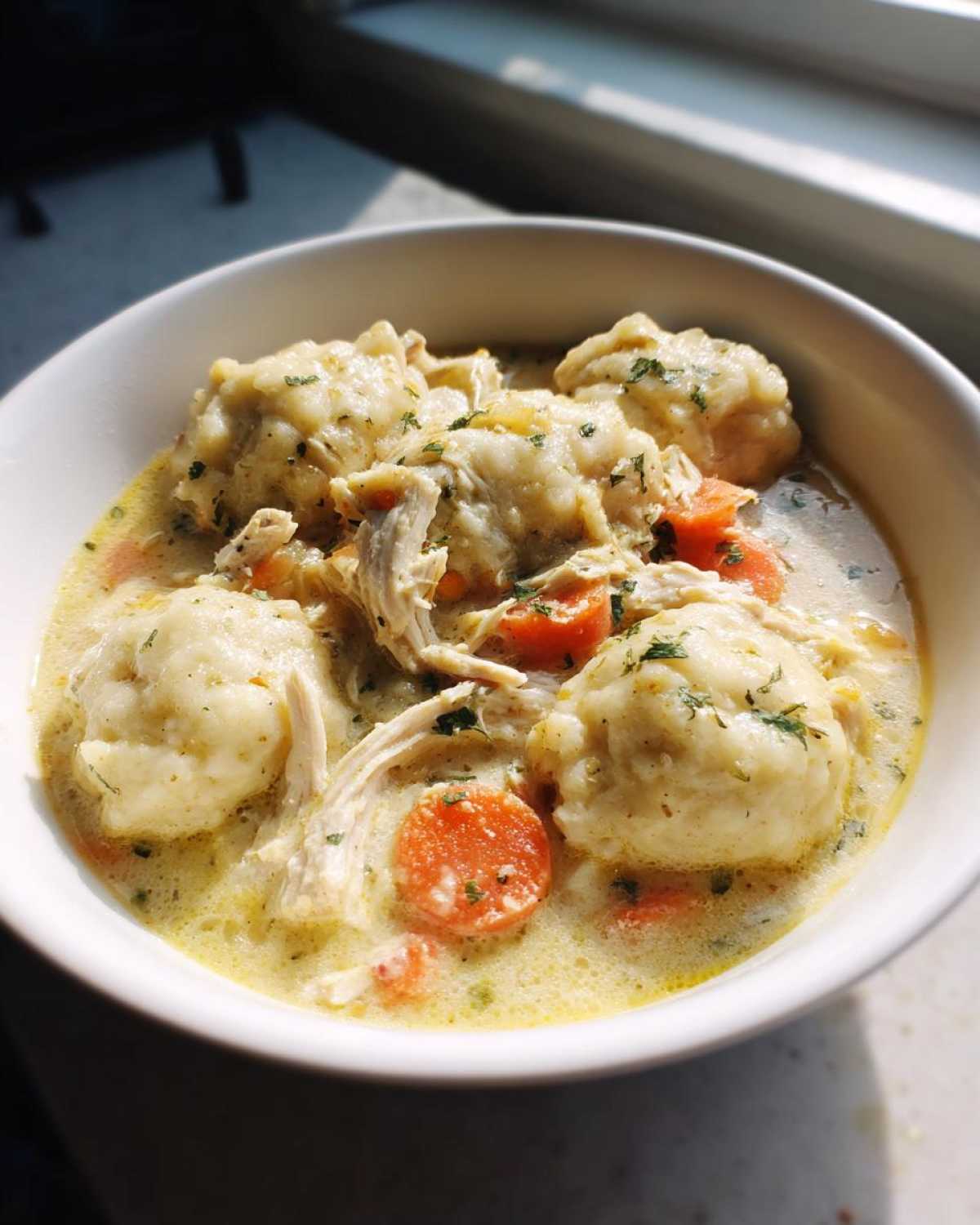 Close-up of a white bowl filled with creamy Instant Pot Chicken and Dumplings, featuring shredded chicken, carrots, and herbs.