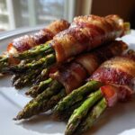 Close-up of perfectly cooked Keto Bacon Wrapped Asparagus Bundles stacked on a white serving plate.
