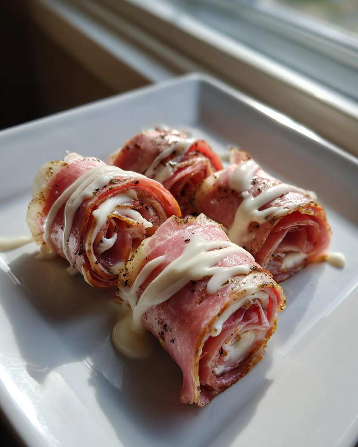 Four Keto Italian Sub Roll-Ups made with deli meat and cheese, drizzled with a white sauce, on a square white plate.