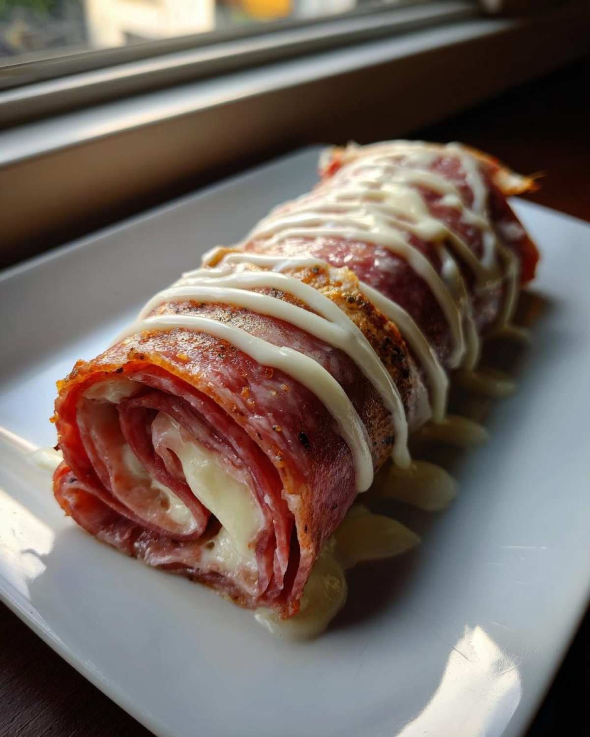 A close-up of Keto Italian Sub Roll-Ups made with salami and melted cheese, drizzled with a white sauce.