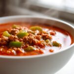 Close-up of a steaming white bowl filled with rich, red Keto Taco Soup with Ground Beef and green bell peppers.