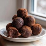 A stack of rich, dark No-Bake Brownie Batter Protein Balls dusted heavily with cocoa powder on a white plate.