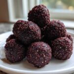 A stack of dark, rich No-Bake Brownie Batter Protein Balls coated in fine crumbs on a white plate.