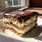A square slice of No-Bake Chocolate Eclair Cake with layers of cream filling and graham crackers, topped with rich chocolate ganache.