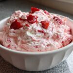 A close-up of fluffy, pink No-Bake Strawberry Cheesecake Dip topped with fresh strawberry pieces in a white bowl.