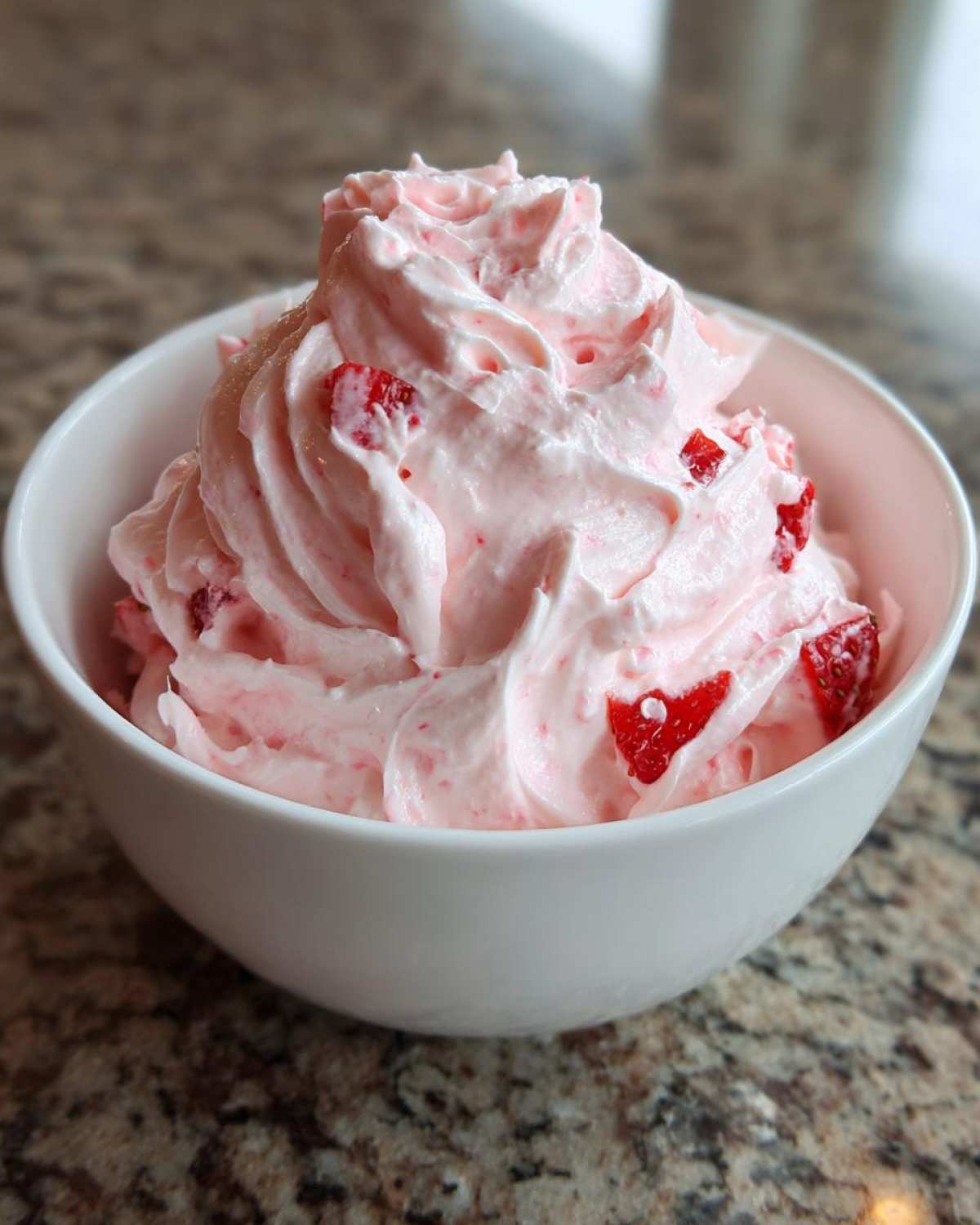 A white bowl filled with fluffy pink No-Bake Strawberry Cheesecake Dip mixed with visible chunks of fresh strawberries.