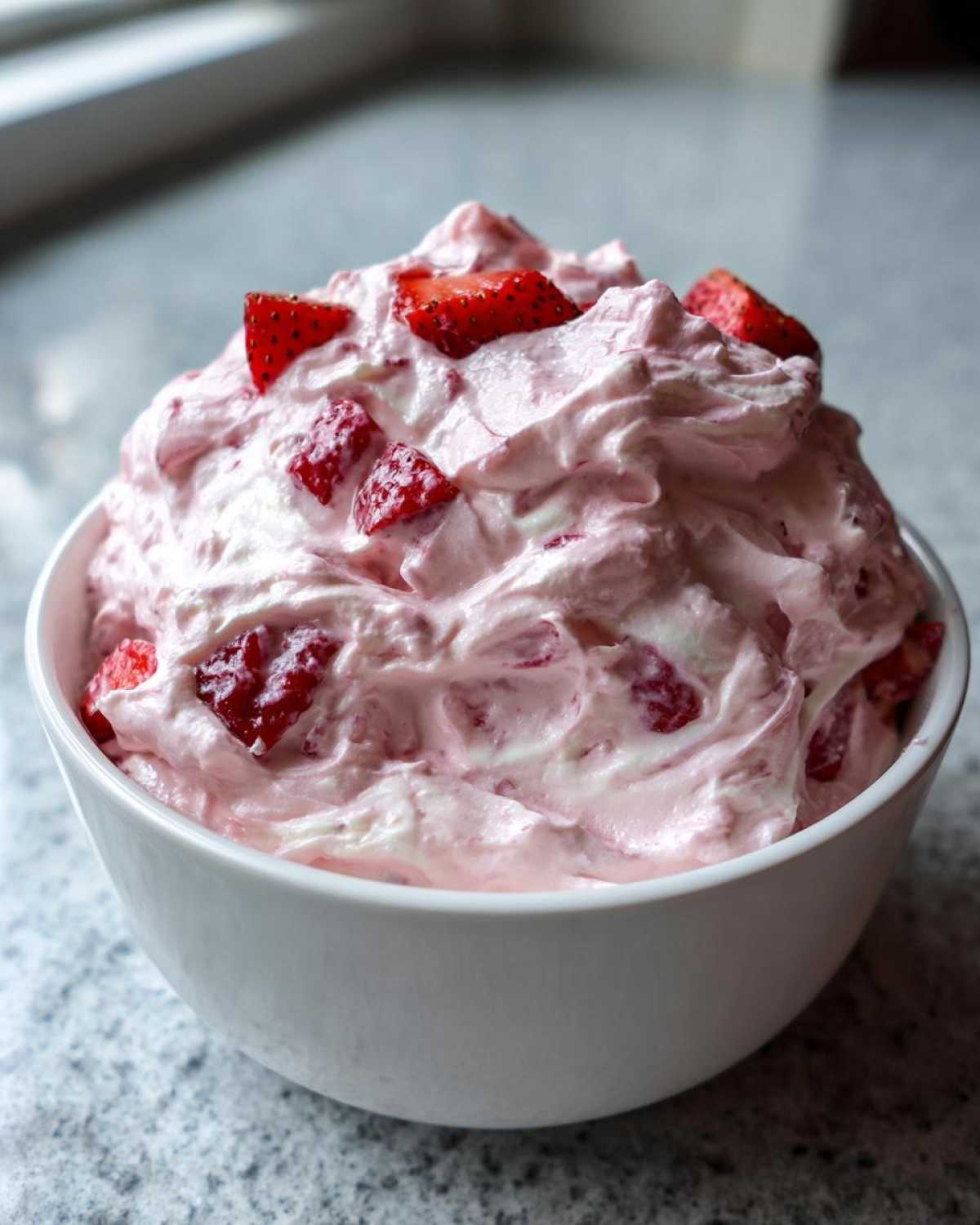 A close-up of fluffy, pink No-Bake Strawberry Cheesecake Dip topped with fresh strawberry chunks in a white bowl.