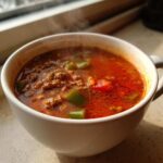 Close-up of a steaming white mug filled with rich, red Keto Taco Soup with Ground Beef and visible green peppers.