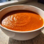 A close-up of a white bowl filled with vibrant orange Vegan Creamy Tomato Coconut Soup.