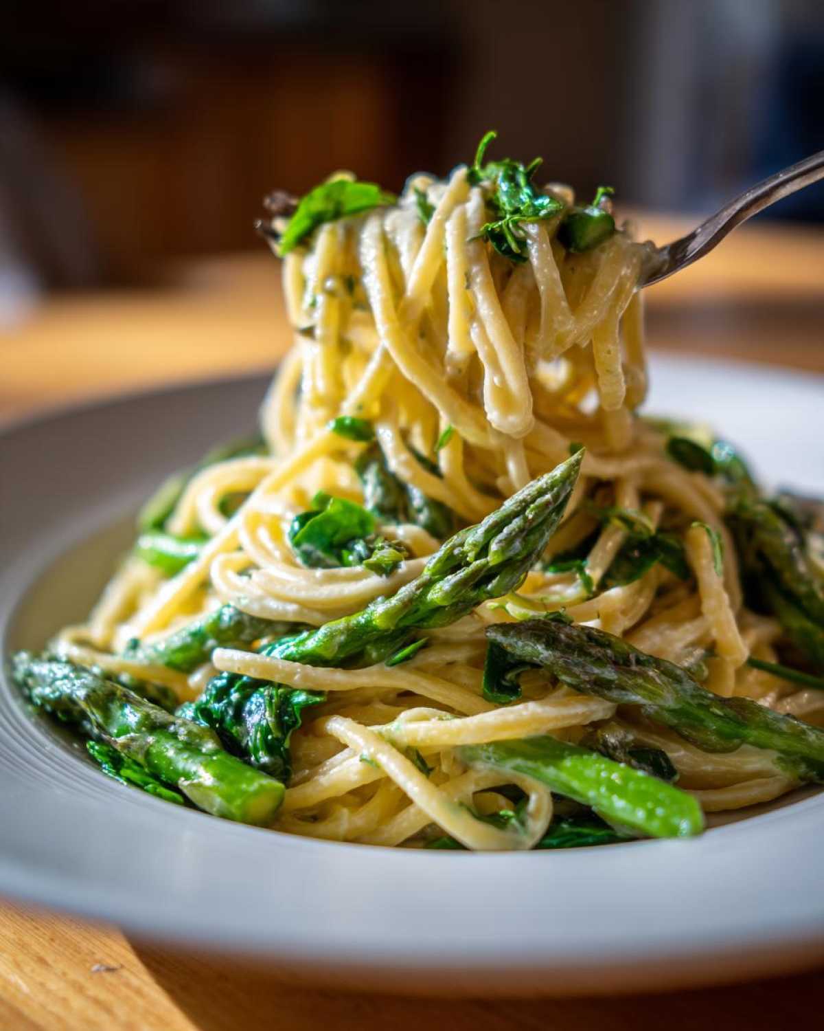 A fork lifts creamy Vegan Lemon Asparagus Pasta with bright green asparagus spears and pea shoots from a white bowl.
