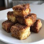 A stack of golden Air Fryer Crispy Cinnamon French Toast Bites coated in cinnamon sugar on a white plate.