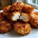 A pile of golden brown Air Fryer Crispy Coconut Chicken Bites on a white plate, one piece broken open showing white chicken inside.