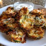 A plate piled high with golden brown Air Fryer Crispy Garlic Dill Pickles, topped with Parmesan, next to a small bowl of white dipping sauce.