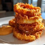 Close-up of golden brown Air Fryer Crispy Onion Petals stacked on a white plate next to a dollop of spicy mayo.