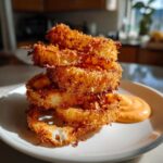 A stack of golden brown, crispy Air Fryer Crispy Onion Petals served on a white plate next to a swirl of spicy mayo.