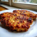 Two golden brown pieces of Air Fryer Crispy Parmesan Pork Schnitzel resting on a white plate near a window.