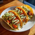 Three crispy tacos filled with orange sweet potato chunks, shredded lettuce, and drizzled with light green sauce, ready to eat.