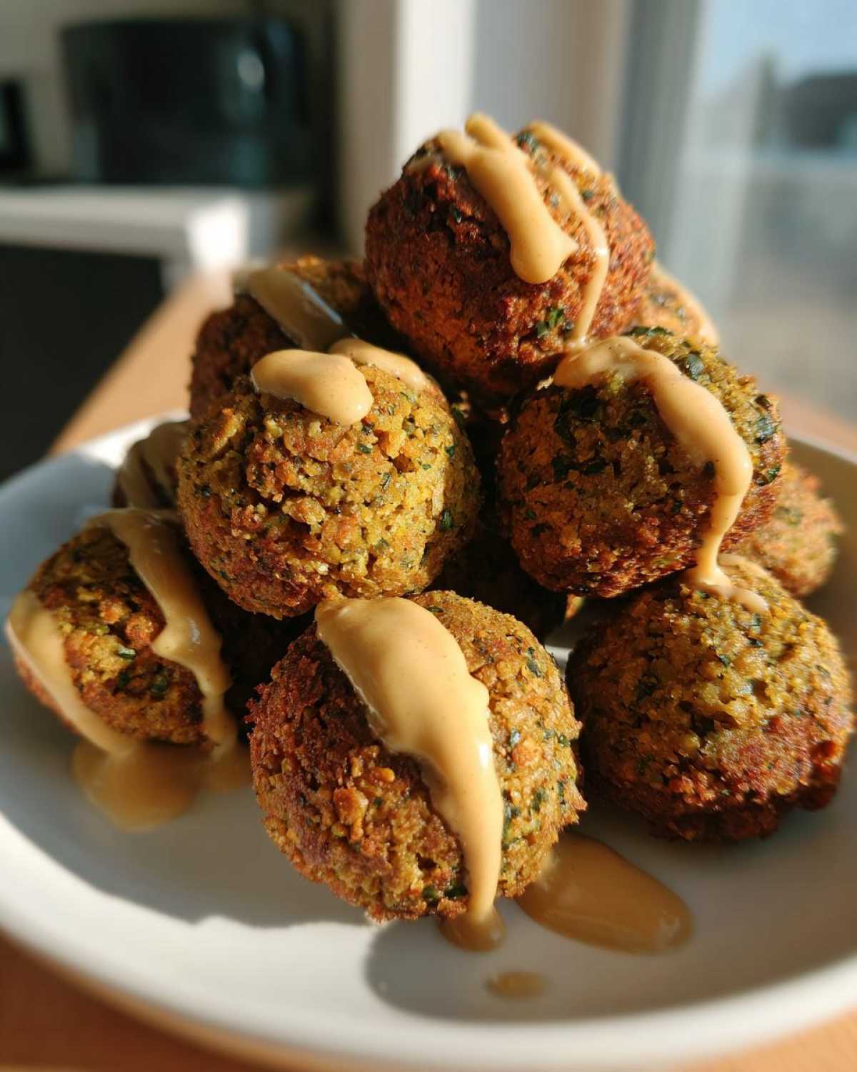 A pile of golden brown Air Fryer Herb Falafel Balls generously drizzled with creamy tahini sauce on a white plate.