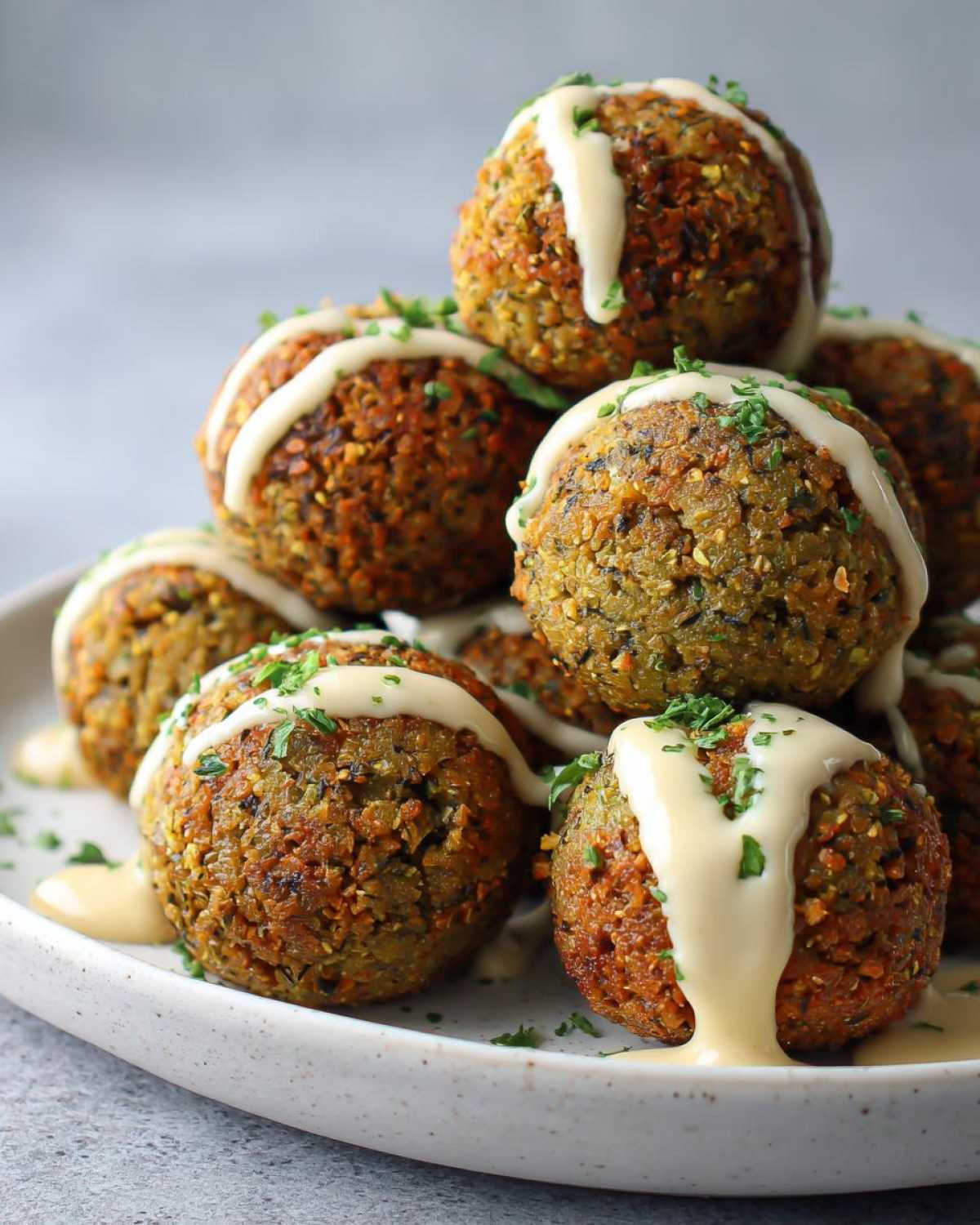 A stack of golden brown Air Fryer Herb Falafel Balls drizzled with creamy tahini sauce and sprinkled with parsley.