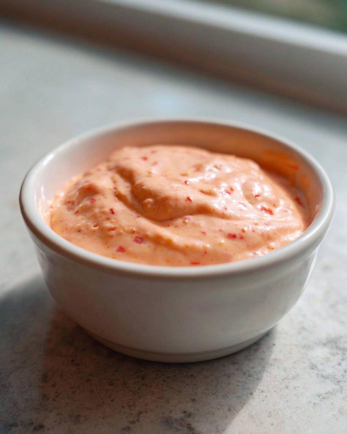 Close-up of a small white bowl filled with creamy, orange-pink Copycat Classic Burger Spread Sauce speckled with red bits.