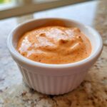 Close-up of creamy, orange-hued Copycat Classic Burger Spread Sauce speckled with red spices in a small white ramekin.