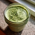 Close-up overhead view of thick, bright green Copycat Green Goddess Ranch Dressing in a glass jar, topped with dried herbs.