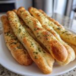 A close-up of several golden Copycat Italian Restaurant Soft Garlic Breadsticks piled on a white plate, topped with garlic butter and parsley.