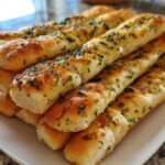 A close-up of several warm, soft garlic breadsticks stacked on a white plate, glistening with butter and herbs.