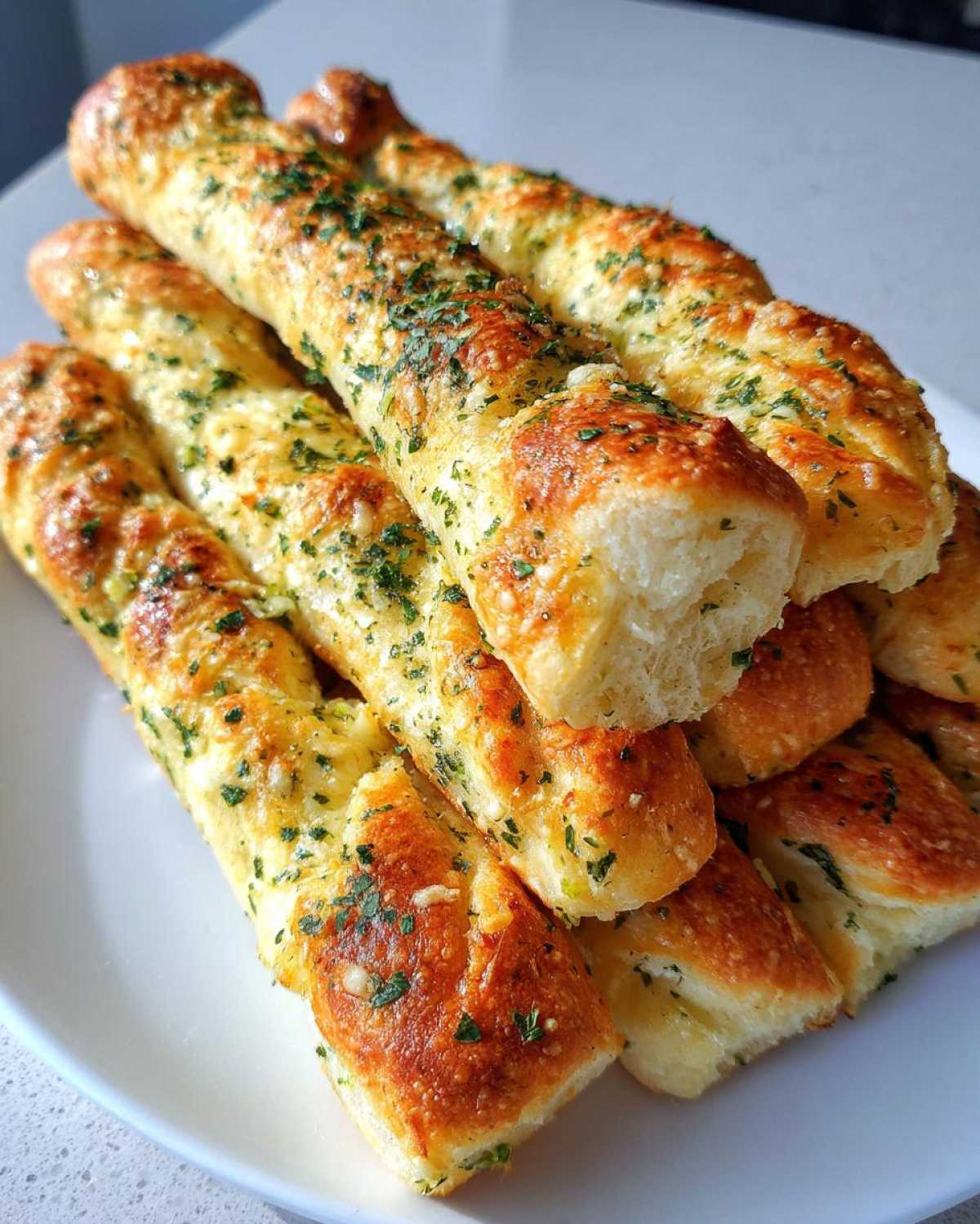 A stack of golden brown Copycat Italian Restaurant Soft Garlic Breadsticks topped with melted butter and parsley on a white plate.