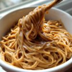 Close-up of noodles being lifted from a white bowl, thoroughly coated in rich, creamy Easy 5-Minute Peanut Sauce.