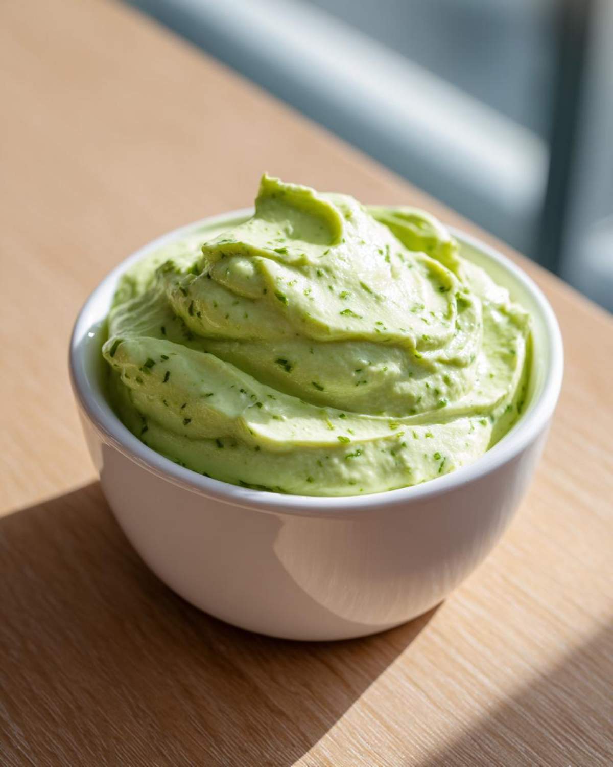 A close-up of a small white bowl filled with thick, pale green Easy 5-Minute Salsa Verde Yogurt Dip, flecked with herbs, sitting on a wooden surface.