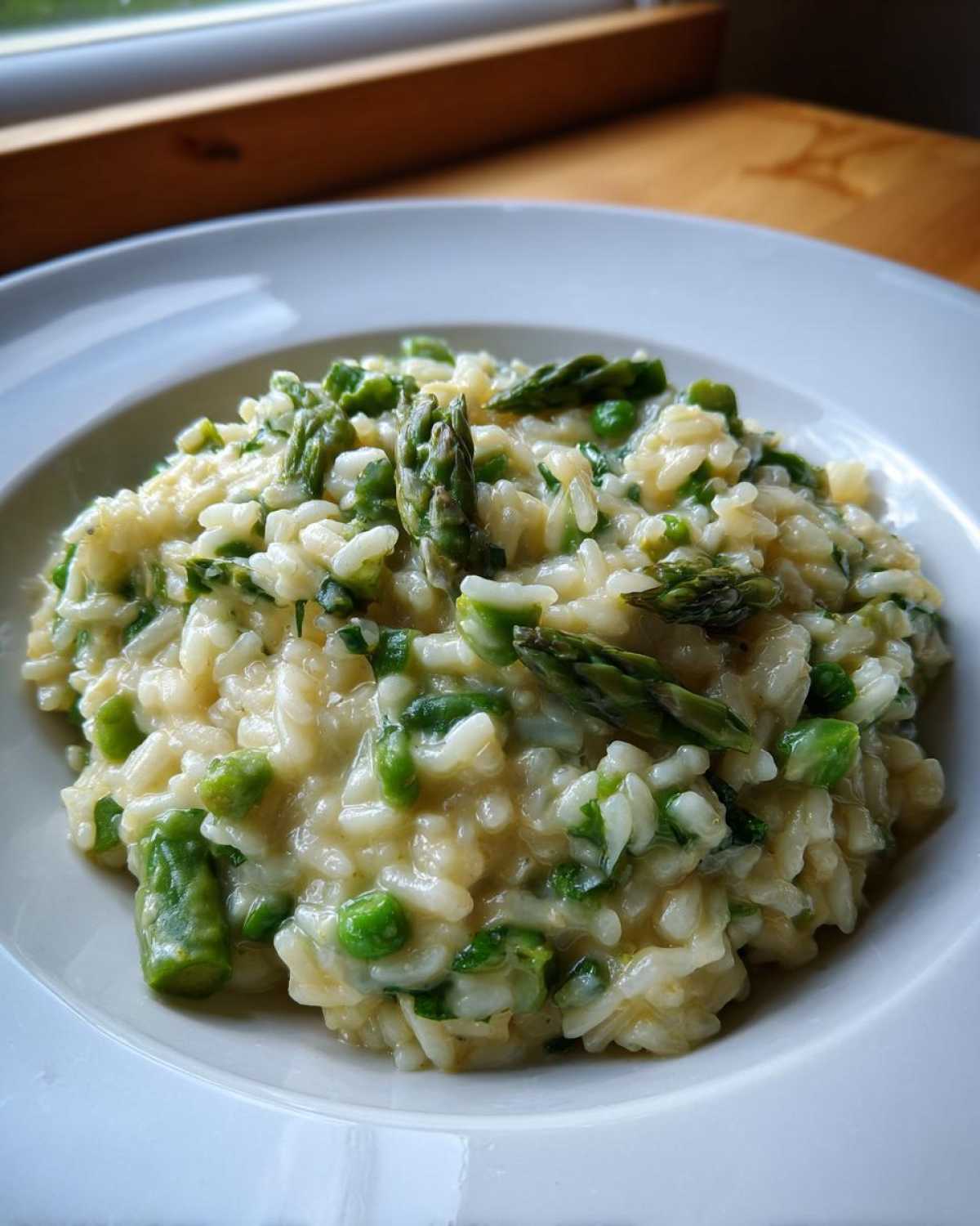 A close-up of a bowl of Easy Creamy Asparagus Risotto mixed with bright green peas and topped with asparagus tips.