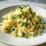 A mound of fluffy, creamy lemon herb couscous garnished generously with fresh chopped parsley on a white plate.