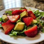 A close-up of a vibrant Easy Strawberry Avocado Salad featuring sliced strawberries, avocado chunks, and mixed greens on a white plate.