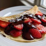 A folded Gluten-Free Chocolate Strawberry Crepes topped with rich chocolate sauce, fresh strawberries, and powdered sugar.