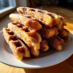 A stack of golden Gluten-Free Cinnamon Sugar Waffle Sticks heavily coated in cinnamon sugar, served on a white plate.
