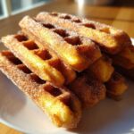 A stack of golden Gluten-Free Cinnamon Sugar Waffle Sticks heavily coated in sparkling cinnamon sugar, resting on a white plate.