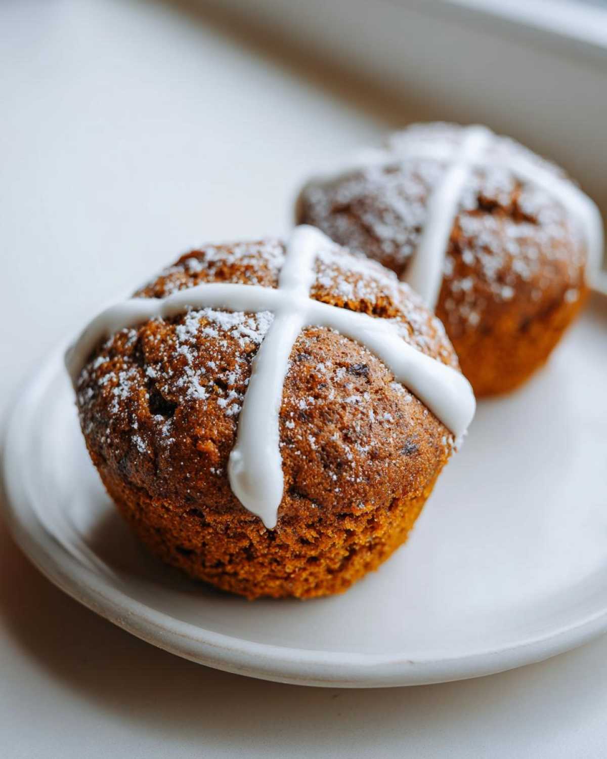 Two delicious Gluten-Free Hot Cross Bun Muffins topped with white icing crosses and powdered sugar on a white plate.