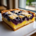 A close-up of a single serving of Gluten-Free Lemon Blueberry Layer Bars showing the crust, lemon filling, and blueberry topping.