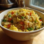A close-up of a white bowl filled with Gluten-Free Lemon Pepper Fried Rice, featuring scrambled egg, peas, and diced carrots.