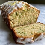 Close-up of Gluten-Free Lemon Zucchini Bread loaf with one slice cut, drizzled with white lemon glaze and zest.