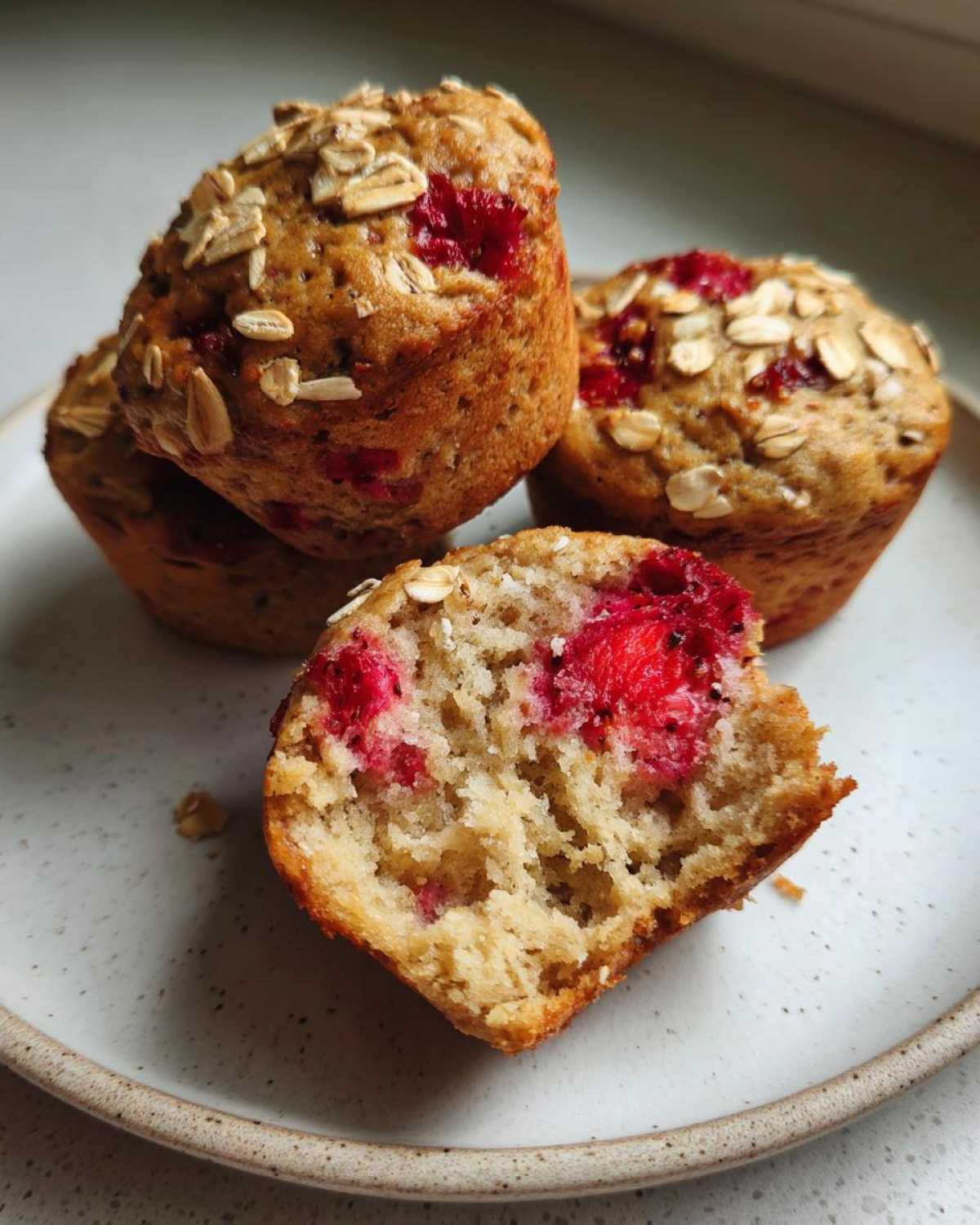 A close-up of several Gluten-Free Strawberry Oat Muffins, one of which is broken open showing the moist interior and bright red strawberries.